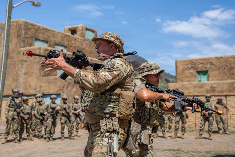 A military training exercise is taking place at what appears to be a mock urban environment or Military Operations on Urban Terrain (MOUT) facility. Two instructors in the foreground are demonstrating weapons handling and tactical movement techniques to a group of soldiers standing in formation in the background. The setting features adobe-style buildings consistent with a training installation designed to simulate Middle Eastern or desert environments.