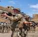 A military training exercise is taking place at what appears to be a mock urban environment or Military Operations on Urban Terrain (MOUT) facility. Two instructors in the foreground are demonstrating weapons handling and tactical movement techniques to a group of soldiers standing in formation in the background. The setting features adobe-style buildings consistent with a training installation designed to simulate Middle Eastern or desert environments.
