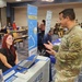 A military service member in US Army OCP (Operational Camouflage Pattern) uniform is conversing with a female booth representative at what appears to be a military resource or benefits fair. Multiple vendor booths are visible including Popular Mortgage, American Red Cross, and Pentagon Federal Credit Union. The event is held in a large conference or banquet hall with multiple attendees and exhibitors in the background.