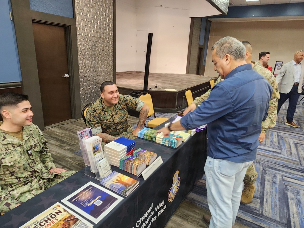 A military chapel resource table is set up in what appears to be a conference or event hall, with service members from multiple branches staffing the booth. Two uniformed personnel — one in Marine Corps MARPAT camouflage and one in Navy NWU (digital blue) uniform — are seated behind a table covered with religious books and pamphlets. A civilian in a blue shirt is interacting with the table, examining the materials on display.