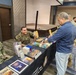 A military chapel resource table is set up in what appears to be a conference or event hall, with service members from multiple branches staffing the booth. Two uniformed personnel — one in Marine Corps MARPAT camouflage and one in Navy NWU (digital blue) uniform — are seated behind a table covered with religious books and pamphlets. A civilian in a blue shirt is interacting with the table, examining the materials on display.