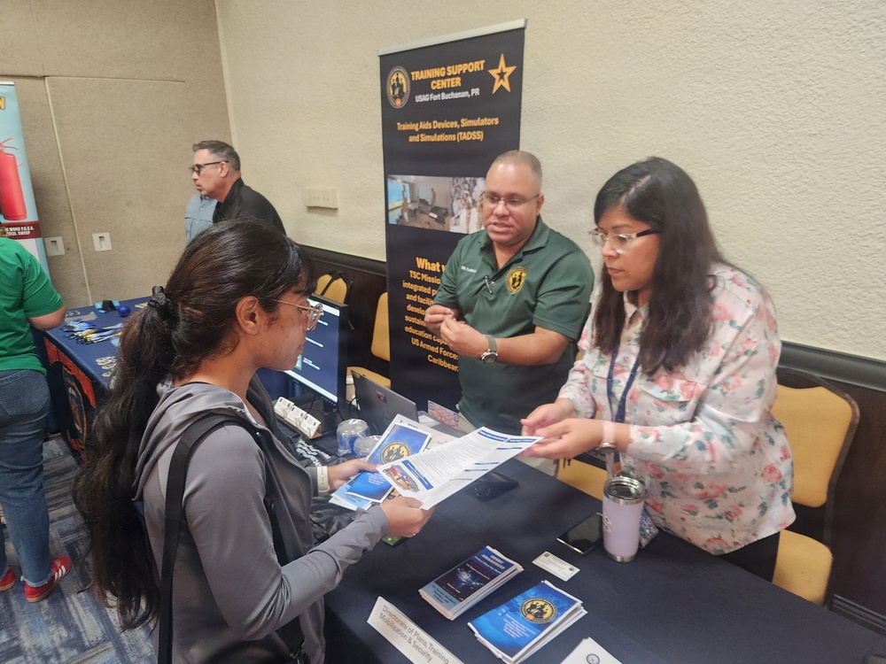 A career or information fair booth representing the Training Support Center (TSC) at USAG Fort Buchanan, Puerto Rico. Two booth representatives are engaging with a visitor, sharing printed brochures and materials about Training Aids Devices, Simulators and Simulations (TADSS). The setting appears to be an indoor conference or event hall.