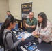 A career or information fair booth representing the Training Support Center (TSC) at USAG Fort Buchanan, Puerto Rico. Two booth representatives are engaging with a visitor, sharing printed brochures and materials about Training Aids Devices, Simulators and Simulations (TADSS). The setting appears to be an indoor conference or event hall.