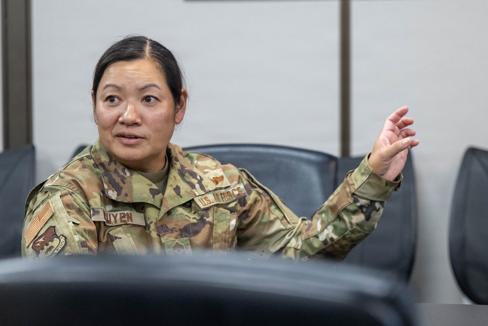 A U.S. Air Force service member wearing OCP (Operational Camouflage Pattern) uniform is seated in what appears to be a briefing room or conference room. The individual, identified by name tape as 'NGUYEN,' is actively speaking and gesturing with one raised hand. The setting suggests a formal military meeting or discussion environment.