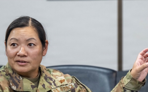 A U.S. Air Force service member wearing OCP (Operational Camouflage Pattern) uniform is seated in what appears to be a briefing room or conference room. The individual, identified by name tape as 'NGUYEN,' is actively speaking and gesturing with one raised hand. The setting suggests a formal military meeting or discussion environment.