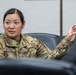 A U.S. Air Force service member wearing OCP (Operational Camouflage Pattern) uniform is seated in what appears to be a briefing room or conference room. The individual, identified by name tape as 'NGUYEN,' is actively speaking and gesturing with one raised hand. The setting suggests a formal military meeting or discussion environment.