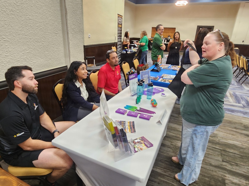 The image shows an indoor event, likely a college or career fair, taking place in a banquet or conference hall. Several booths are set up with promotional materials, and representatives are engaging with visitors. Multiple organizations appear to be represented, with staff seated at tables interacting with attendees.