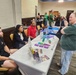 The image shows an indoor event, likely a college or career fair, taking place in a banquet or conference hall. Several booths are set up with promotional materials, and representatives are engaging with visitors. Multiple organizations appear to be represented, with staff seated at tables interacting with attendees.