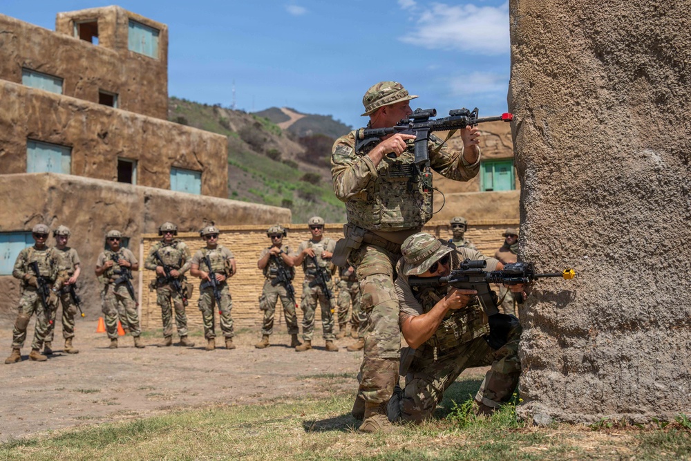 Military personnel are conducting a tactical training exercise at what appears to be a mock urban environment or Military Operations on Urban Terrain (MOUT) facility. Two instructors or demonstrators are positioned at a wall corner, one standing and one kneeling, both aiming rifles in a stacked formation. A group of approximately 12-15 soldiers stands in the background observing the demonstration.