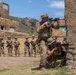Military personnel are conducting a tactical training exercise at what appears to be a mock urban environment or Military Operations on Urban Terrain (MOUT) facility. Two instructors or demonstrators are positioned at a wall corner, one standing and one kneeling, both aiming rifles in a stacked formation. A group of approximately 12-15 soldiers stands in the background observing the demonstration.