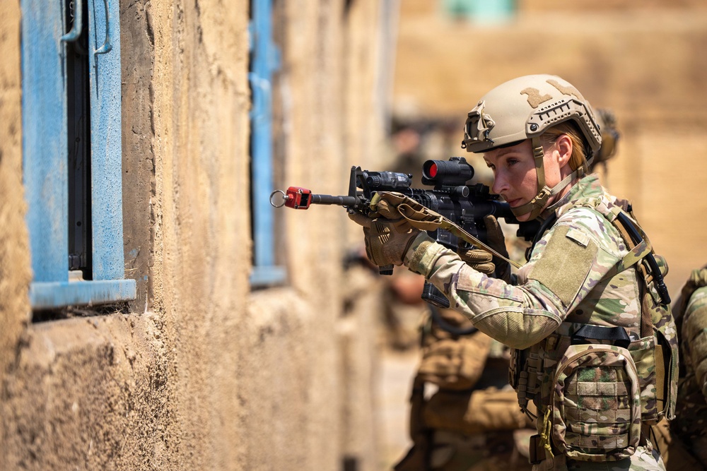 A female soldier in full combat gear is conducting a tactical building clearance exercise, aiming a rifle toward a window on a tan concrete structure. She is wearing MultiCam/OCP camouflage uniform, a tan tactical helmet, and full plate carrier with pouches. Additional personnel are visible in the blurred background, suggesting a team-based military training or operational scenario.