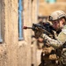 A female soldier in full combat gear is conducting a tactical building clearance exercise, aiming a rifle toward a window on a tan concrete structure. She is wearing MultiCam/OCP camouflage uniform, a tan tactical helmet, and full plate carrier with pouches. Additional personnel are visible in the blurred background, suggesting a team-based military training or operational scenario.