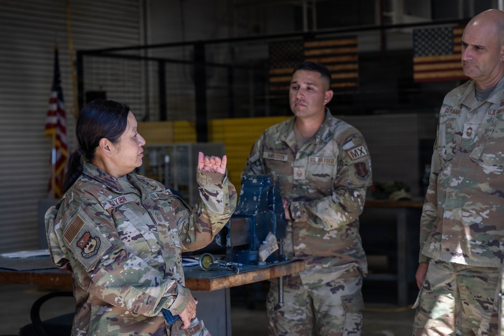 Three U.S. Air Force personnel in OCP (Operational Camouflage Pattern) uniforms are engaged in a discussion inside what appears to be a maintenance hangar or workshop facility. A female airman on the left is speaking and gesturing with her hands while two male airmen listen attentively. American flags are visible in the background, and a workbench with equipment is present.