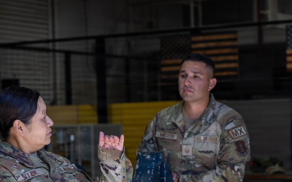 Three U.S. Air Force personnel in OCP (Operational Camouflage Pattern) uniforms are engaged in a discussion inside what appears to be a maintenance hangar or workshop facility. A female airman on the left is speaking and gesturing with her hands while two male airmen listen attentively. American flags are visible in the background, and a workbench with equipment is present.