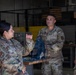 Three U.S. Air Force personnel in OCP (Operational Camouflage Pattern) uniforms are engaged in a discussion inside what appears to be a maintenance hangar or workshop facility. A female airman on the left is speaking and gesturing with her hands while two male airmen listen attentively. American flags are visible in the background, and a workbench with equipment is present.