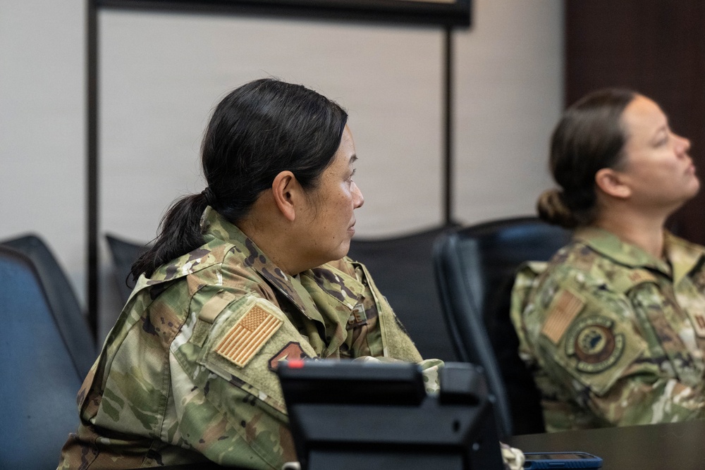 Two female military personnel in OCP (Operational Camouflage Pattern) uniforms are seated at what appears to be a briefing or training room. Both wear U.S. flag patches on their right shoulders, indicating active U.S. military service. They appear to be attentively observing a presentation or briefing taking place off-camera.