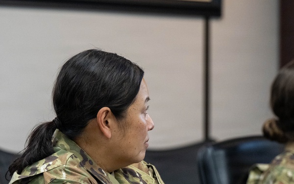 Two female military personnel in OCP (Operational Camouflage Pattern) uniforms are seated at what appears to be a briefing or training room. Both wear U.S. flag patches on their right shoulders, indicating active U.S. military service. They appear to be attentively observing a presentation or briefing taking place off-camera.