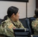 Two female military personnel in OCP (Operational Camouflage Pattern) uniforms are seated at what appears to be a briefing or training room. Both wear U.S. flag patches on their right shoulders, indicating active U.S. military service. They appear to be attentively observing a presentation or briefing taking place off-camera.