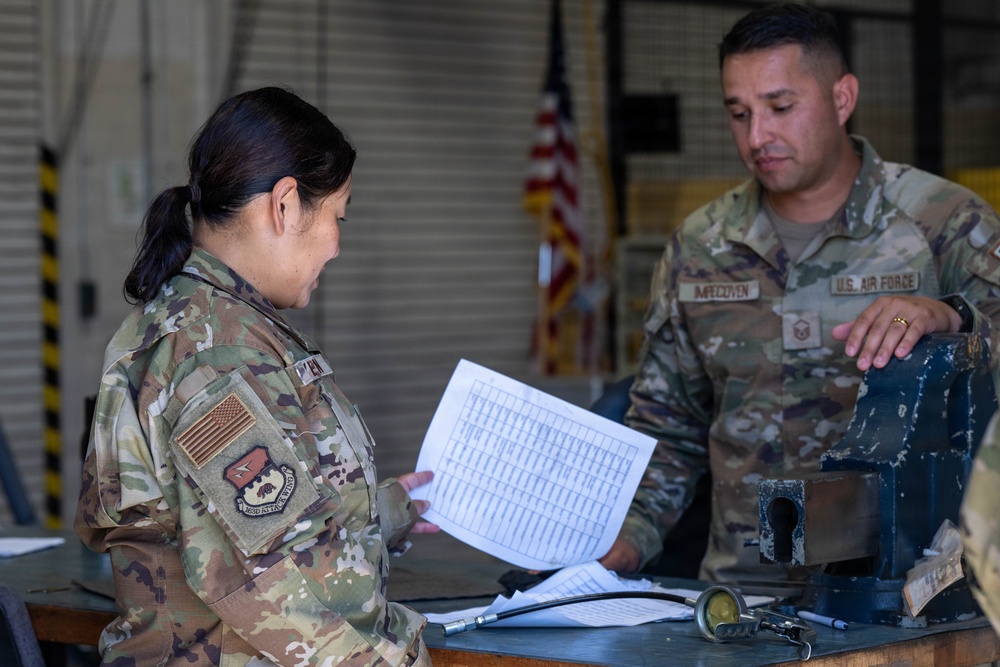 Two U.S. Air Force personnel in OCP (Operational Camouflage Pattern) uniforms are reviewing a document together in what appears to be a maintenance hangar or workshop. The male airman, whose name tape reads 'IMPECOVEN,' is pointing at or handling equipment on a workbench while the female airman reviews a printed checklist or data sheet. An American flag is visible in the background.
