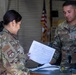 Two U.S. Air Force personnel in OCP (Operational Camouflage Pattern) uniforms are reviewing a document together in what appears to be a maintenance hangar or workshop. The male airman, whose name tape reads 'IMPECOVEN,' is pointing at or handling equipment on a workbench while the female airman reviews a printed checklist or data sheet. An American flag is visible in the background.