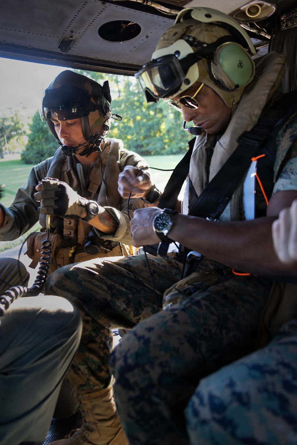 Two military personnel wearing flight helmets with communication equipment are seated inside what appears to be a military helicopter. They appear to be engaged in a medical or technical procedure, with one individual attending to another person's limb. The setting suggests a medevac or training exercise scenario.