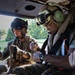 Two military personnel wearing flight helmets with communication equipment are seated inside what appears to be a military helicopter. They appear to be engaged in a medical or technical procedure, with one individual attending to another person's limb. The setting suggests a medevac or training exercise scenario.