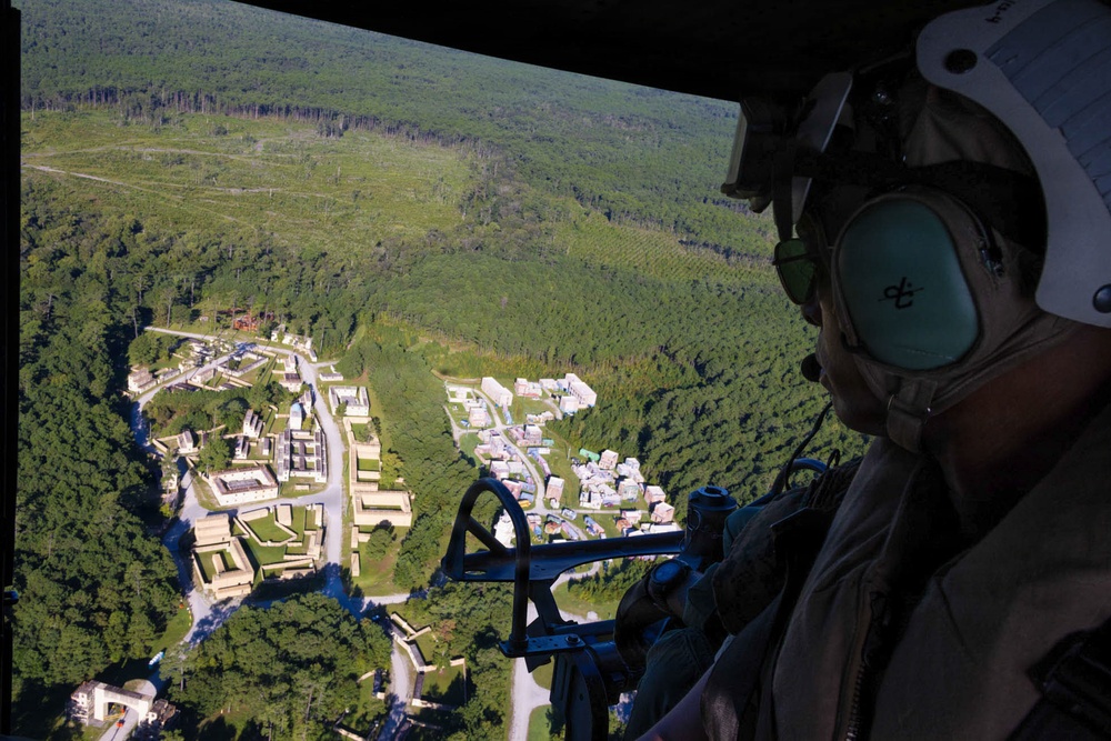 An aerial photograph taken from inside a military helicopter, showing a crew member in flight gear observing a simulated urban training village below. The settlement is surrounded by dense forest and features numerous small structures arranged along roads, consistent with a military combat training facility. The perspective is from the open door or window of the helicopter looking outward and downward.