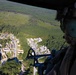 An aerial photograph taken from inside a military helicopter, showing a crew member in flight gear observing a simulated urban training village below. The settlement is surrounded by dense forest and features numerous small structures arranged along roads, consistent with a military combat training facility. The perspective is from the open door or window of the helicopter looking outward and downward.
