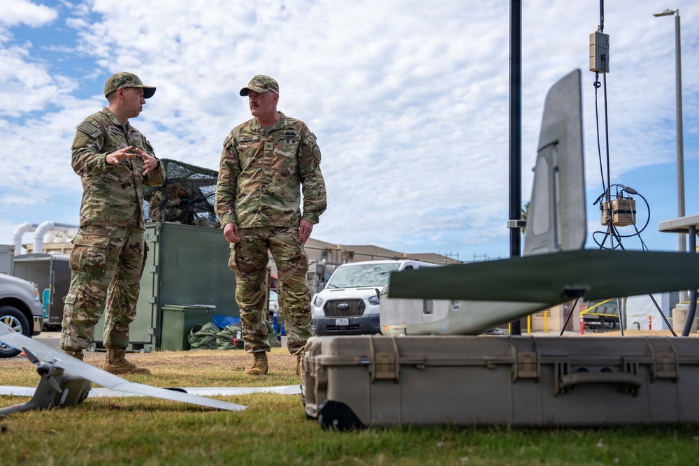 Two military personnel in OCP (Operational Camouflage Pattern) uniforms are engaged in conversation at what appears to be an outdoor military equipment demonstration or training area. In the foreground, unmanned aerial vehicle (UAV/drone) components and military transport cases are visible, along with communication antenna equipment. The setting appears to be a military installation with vehicles and structures visible in the background.