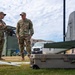 Two military personnel in OCP (Operational Camouflage Pattern) uniforms are engaged in conversation at what appears to be an outdoor military equipment demonstration or training area. In the foreground, unmanned aerial vehicle (UAV/drone) components and military transport cases are visible, along with communication antenna equipment. The setting appears to be a military installation with vehicles and structures visible in the background.