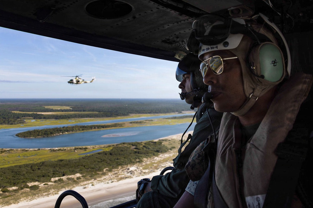 Two military aircrew members are photographed from inside a helicopter in flight, wearing flight helmets, communication headsets, and aviator sunglasses. A second military helicopter, appearing to be a Bell UH-1Y Venom or similar utility helicopter, is visible flying in formation in the background. Below, a coastal landscape featuring a river estuary, sandy beach, marshland, and forested terrain is visible.