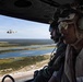 Two military aircrew members are photographed from inside a helicopter in flight, wearing flight helmets, communication headsets, and aviator sunglasses. A second military helicopter, appearing to be a Bell UH-1Y Venom or similar utility helicopter, is visible flying in formation in the background. Below, a coastal landscape featuring a river estuary, sandy beach, marshland, and forested terrain is visible.
