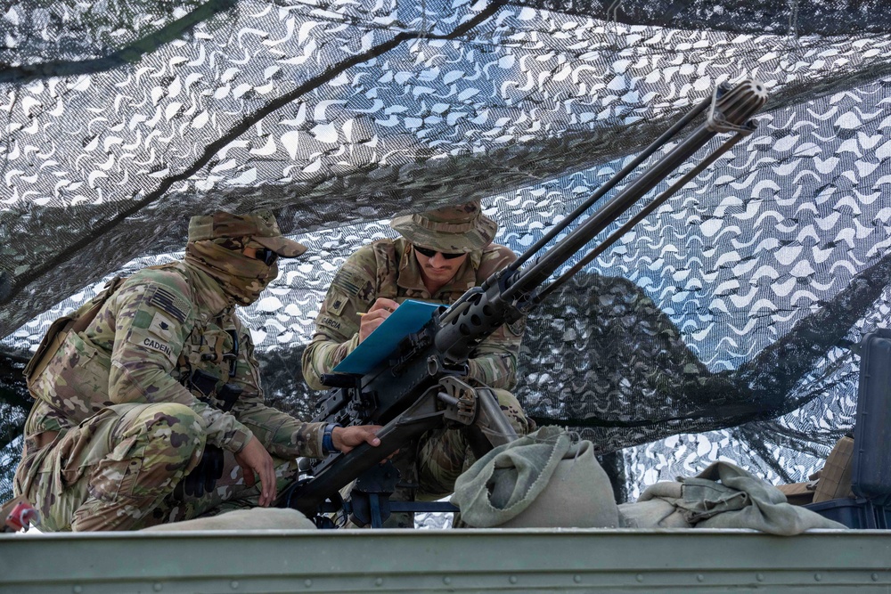 Two U.S. Army soldiers in Operational Camouflage Pattern (OCP) uniforms are positioned under a camouflage net, reviewing a document on a blue clipboard near a mounted heavy machine gun, likely an M2 .50-caliber. The name tapes visible on their uniforms read 'CADEN' (left) and 'GARCIA' (right). The setting appears to be a field training exercise or tactical position on or near a military vehicle.