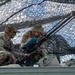 Two U.S. Army soldiers in Operational Camouflage Pattern (OCP) uniforms are positioned under a camouflage net, reviewing a document on a blue clipboard near a mounted heavy machine gun, likely an M2 .50-caliber. The name tapes visible on their uniforms read 'CADEN' (left) and 'GARCIA' (right). The setting appears to be a field training exercise or tactical position on or near a military vehicle.