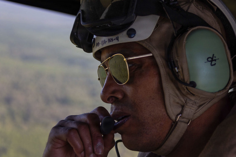 A military aviator or crew member is photographed in close-up while airborne, wearing a flight helmet with communications headset and aviator-style sunglasses. The individual is holding a microphone or push-to-talk device near their mouth, suggesting active radio communication. A green, vegetated landscape is visible through an open aircraft door or window in the background.