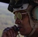 A military aviator or crew member is photographed in close-up while airborne, wearing a flight helmet with communications headset and aviator-style sunglasses. The individual is holding a microphone or push-to-talk device near their mouth, suggesting active radio communication. A green, vegetated landscape is visible through an open aircraft door or window in the background.
