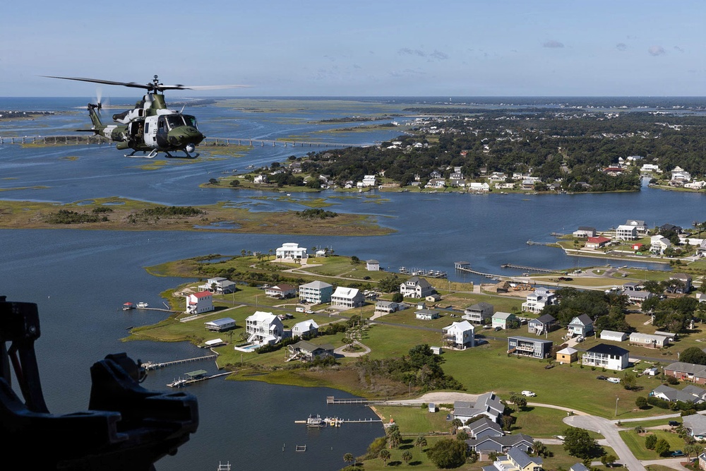An aerial photograph taken from one helicopter showing a second military helicopter flying over a coastal waterway community. The scene depicts a low-altitude flight over a network of islands, marshlands, and residential neighborhoods with waterfront properties. The landscape appears to be a southeastern U.S. coastal estuary region with numerous inlets, docks, and scattered housing developments.