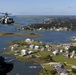 An aerial photograph taken from one helicopter showing a second military helicopter flying over a coastal waterway community. The scene depicts a low-altitude flight over a network of islands, marshlands, and residential neighborhoods with waterfront properties. The landscape appears to be a southeastern U.S. coastal estuary region with numerous inlets, docks, and scattered housing developments.