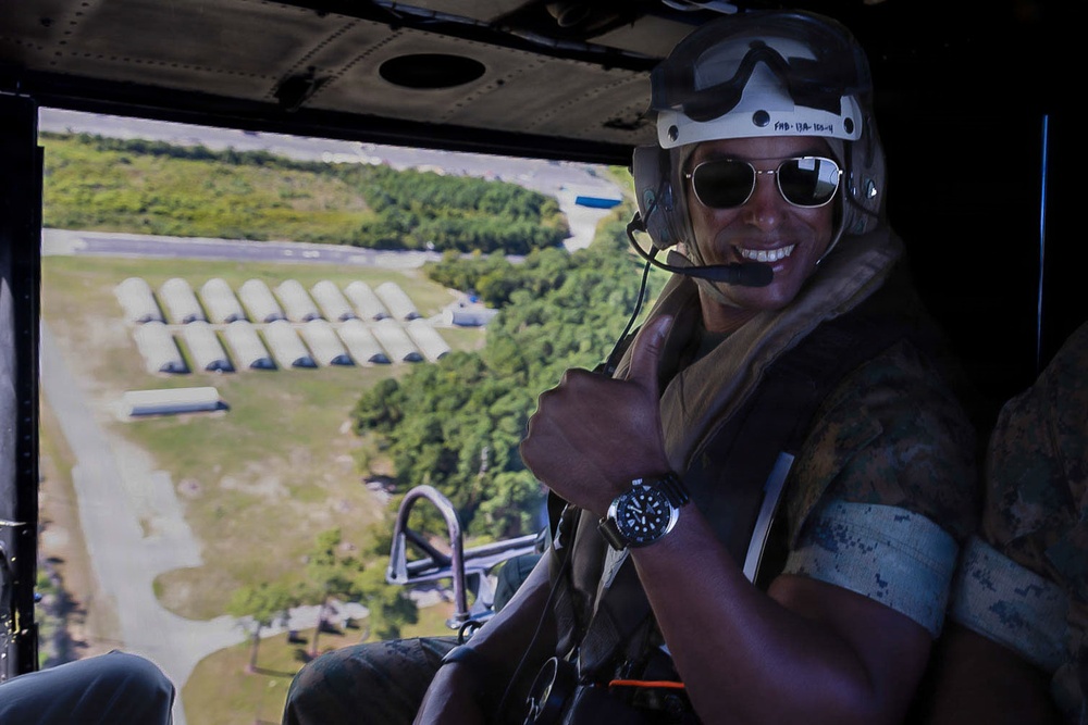A smiling military personnel is seated inside a helicopter in flight, giving a thumbs-up gesture. Below, a military installation or airfield is visible with rows of Quonset hut-style structures and a runway or taxiway. The individual is wearing full flight gear including a helmet, goggles, sunglasses, a communications headset, and camouflage uniform.