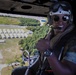 A smiling military personnel is seated inside a helicopter in flight, giving a thumbs-up gesture. Below, a military installation or airfield is visible with rows of Quonset hut-style structures and a runway or taxiway. The individual is wearing full flight gear including a helmet, goggles, sunglasses, a communications headset, and camouflage uniform.