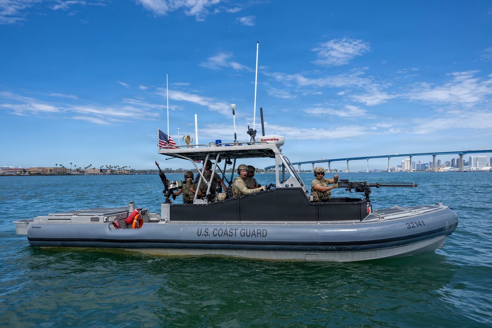 A U.S. Coast Guard rigid-hull inflatable boat (RHIB) numbered 32141 is underway on open water with a crew of armed personnel in military/tactical gear. The vessel is equipped with mounted weapons, navigation equipment including a Raymarine radar unit, and displays an American flag. A large bridge and urban skyline are visible in the background, consistent with San Diego Bay and the Coronado Bridge.