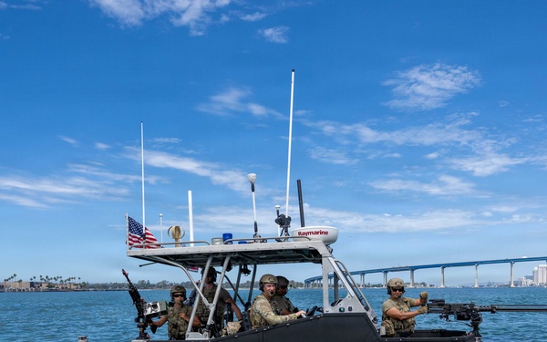 A U.S. Coast Guard rigid-hull inflatable boat (RHIB) numbered 32141 is underway on open water with a crew of armed personnel in military/tactical gear. The vessel is equipped with mounted weapons, navigation equipment including a Raymarine radar unit, and displays an American flag. A large bridge and urban skyline are visible in the background, consistent with San Diego Bay and the Coronado Bridge.