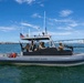 A U.S. Coast Guard rigid-hull inflatable boat (RHIB) numbered 32141 is underway on open water with a crew of armed personnel in military/tactical gear. The vessel is equipped with mounted weapons, navigation equipment including a Raymarine radar unit, and displays an American flag. A large bridge and urban skyline are visible in the background, consistent with San Diego Bay and the Coronado Bridge.