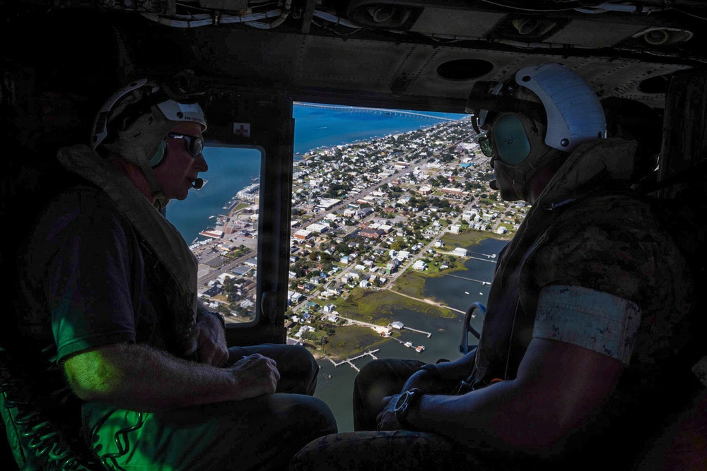 Two military personnel are seated inside a helicopter, observing a coastal town from an aerial vantage point. The view through the open helicopter door reveals a low-lying coastal community with visible flooding or waterlogged areas, a bridge, and a bay or inlet. The mission appears to be a reconnaissance or damage assessment flight, likely following a natural disaster such as a hurricane.