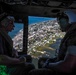 Two military personnel are seated inside a helicopter, observing a coastal town from an aerial vantage point. The view through the open helicopter door reveals a low-lying coastal community with visible flooding or waterlogged areas, a bridge, and a bay or inlet. The mission appears to be a reconnaissance or damage assessment flight, likely following a natural disaster such as a hurricane.