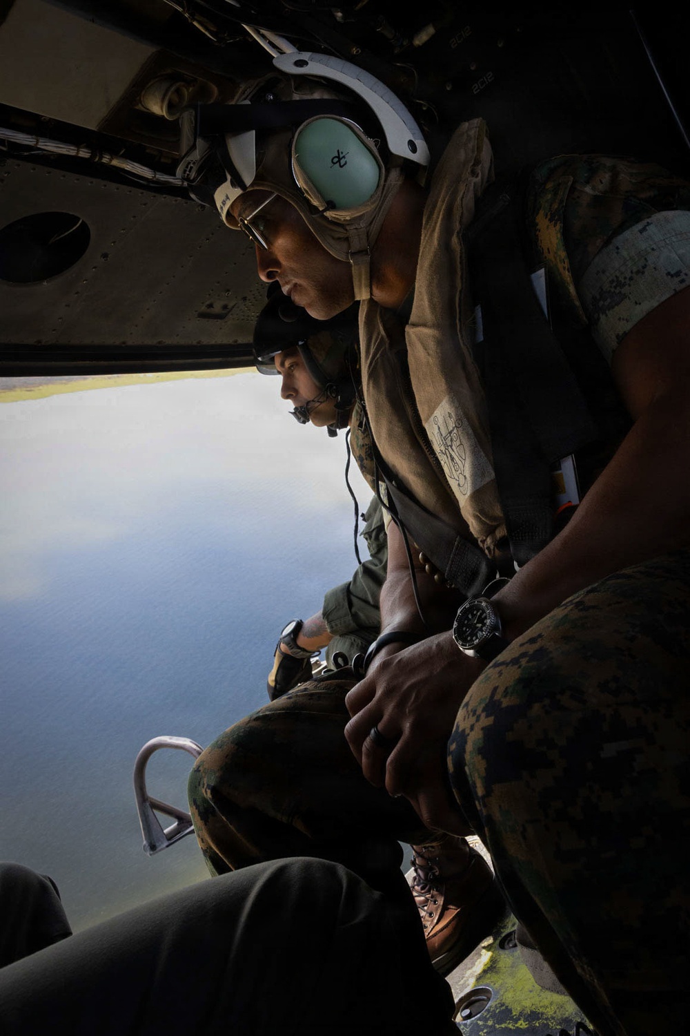 Two military personnel in flight gear are seated inside an open helicopter, looking out over a body of water below. Both individuals are wearing helmets with hearing protection, camouflage uniforms, and tactical vests, consistent with U.S. Marine Corps equipment. The photograph is taken from inside the aircraft, capturing the open door with the aerial view of water and coastline visible beneath them.