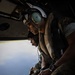 Two military personnel in flight gear are seated inside an open helicopter, looking out over a body of water below. Both individuals are wearing helmets with hearing protection, camouflage uniforms, and tactical vests, consistent with U.S. Marine Corps equipment. The photograph is taken from inside the aircraft, capturing the open door with the aerial view of water and coastline visible beneath them.