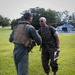 Two military personnel are conversing on a grass field, with one wearing a flight suit and tactical vest and the other in Marine Corps MARPAT camouflage uniform. Aviation helmets and gear are laid on the ground between them. A military helicopter, appearing to be a UH-1Y Venom marked '65', is visible on a landing pad in the background.