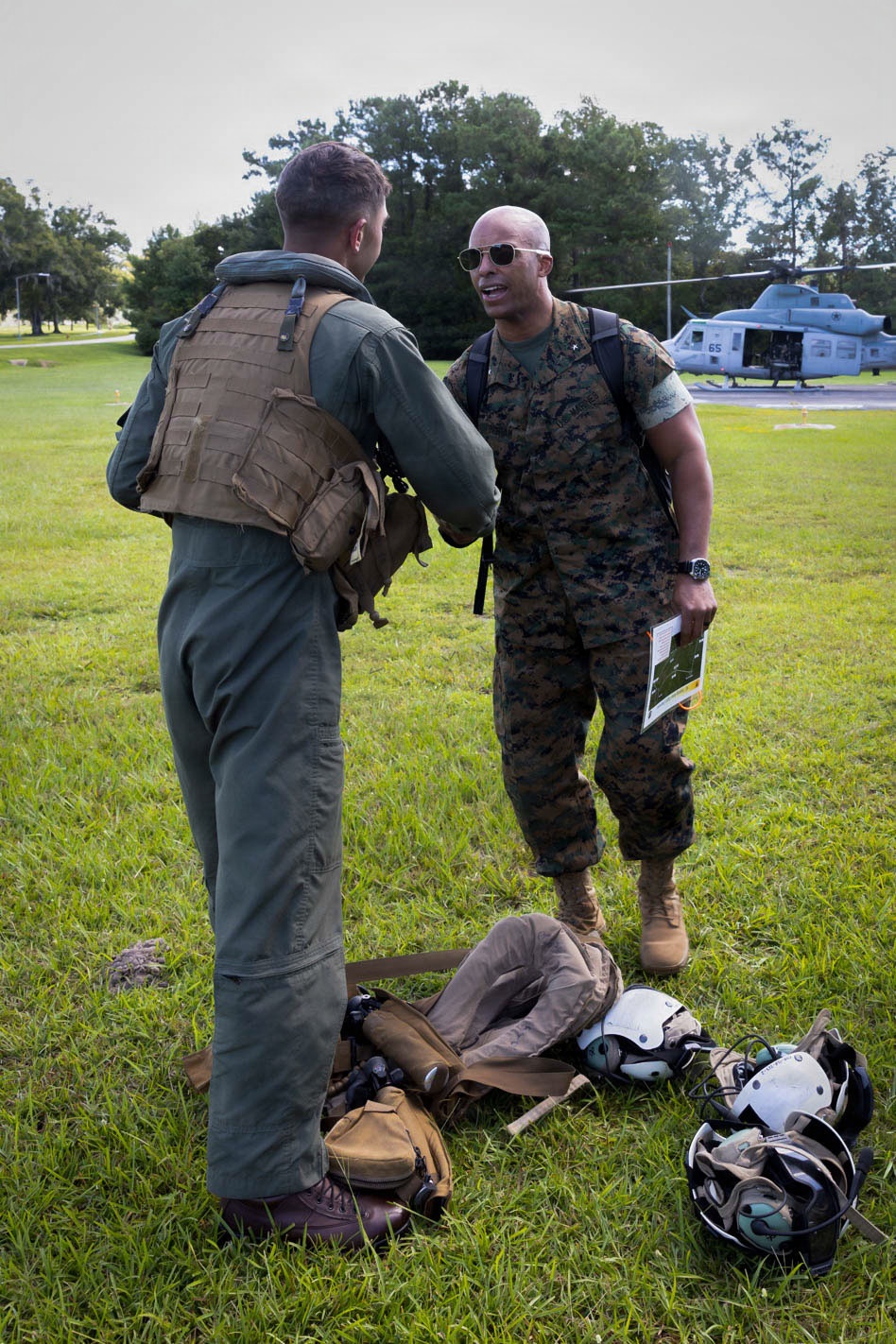Two military personnel are conversing on a grass field, with one wearing a flight suit and tactical vest and the other in Marine Corps MARPAT camouflage uniform. Aviation helmets and gear are laid on the ground between them. A military helicopter, appearing to be a UH-1Y Venom marked '65', is visible on a landing pad in the background.