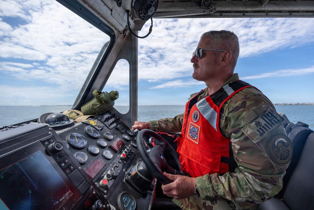 A military or Coast Guard personnel member is piloting a patrol boat on open water. The individual is wearing camouflage fatigues and an orange Coast Guard-marked life vest, operating the vessel from a helm console equipped with multiple navigation instruments. The scene appears to be a routine maritime patrol or operational mission under partly cloudy skies.
